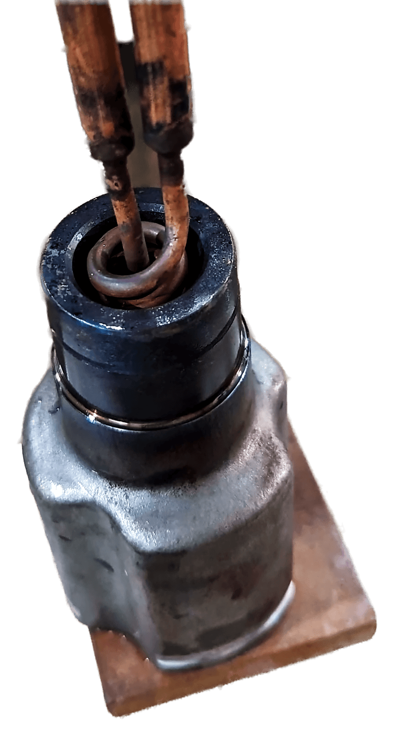 A dark cylindrical steel component sitting inside a copper induction coil, with the coil leads rising vertically out of the machine.
