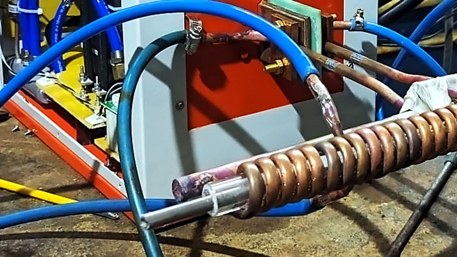 Two hands forming a copper tube induction coil on a bench, with blue cooling hoses and the machine frame visible behind.